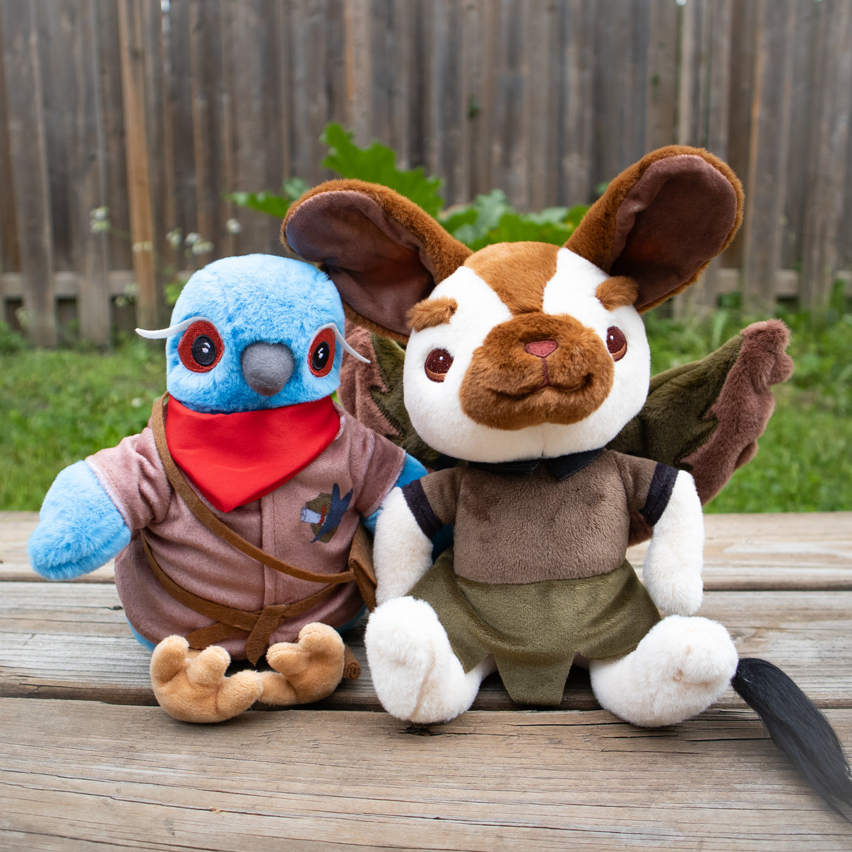 A photo of 2 Humblewood plushies on a wooden porch, outside.